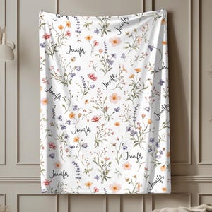 Puede incluir: Manta blanca con un estampado floral en tonos rosa, naranja y morado. El nombre "Jennifer" está escrito en cursiva sobre la manta. La manta está decorada con flores y follaje estilo acuarela.