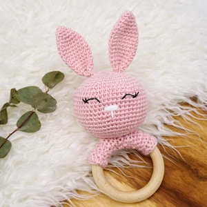 May include: A pink crochet bunny rattle with a wooden ring base. The bunny features long ears, closed eyes, and a small white nose. The baby rattle is set against a white fluffy background.