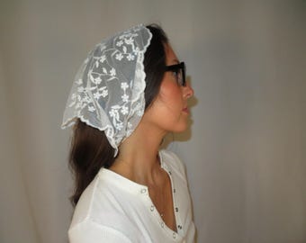 May include: A white lace headscarf with a floral pattern. The head covering is worn on the head. The person is wearing a white shirt and black glasses. The headscarf is a vintage style.