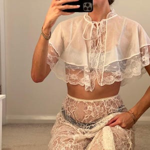Op de afbeelding: Een witte kanten crop top met een hoge halslijn en striksluiting, gecombineerd met een bijpassende kanten rok. De outfit heeft delicate kanten details en een gelaagd ontwerp, wat een romantische en elegante uitstraling creëert.