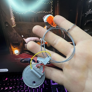 May include: A small, silver-colored, circular component with attached wires and connectors. The wires are gray, yellow, and red. The component is held in a hand, with a ring on one finger.