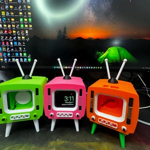 May include: Three colorful, retro-style TV-shaped stands. The stands are green, pink, and orange, each with white legs and antenna. The pink stand displays a digital clock showing the time and alarm.