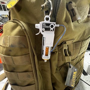 May include: A white and silver keychain resembling a miniature device, attached to a khaki backpack. The device has a trigger, a clear cylinder with an orange coil, and a blue wire. The backpack has multiple pockets and straps.