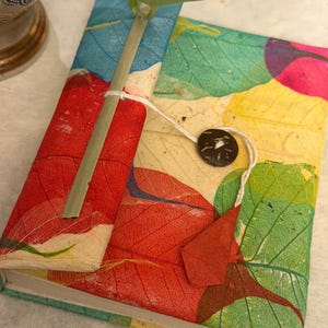 May include: A handmade journal with a cover featuring colorful leaf patterns in red, green, yellow, and blue. The journal is tied with a white string and a dark brown button. A green stem and leaves are placed on top.