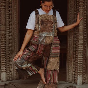 May include: Brown patchwork overalls with a white t-shirt. The overalls feature a variety of patterned fabrics in shades of brown, red, and beige. The garment has a bib front with pockets and wide legs. The overall is worn by a person.