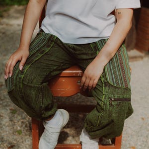 May include: Green patterned pants with a relaxed fit, featuring a side pocket with a zipper. The pants have a unique design with colorful accents. Paired with a white t-shirt and white sneakers, the pants are worn while seated on a wooden chair.