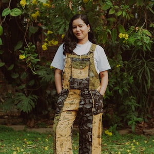 May include: Overalls with a patchwork design featuring mushroom and butterfly illustrations in shades of tan, olive green, and black. The overalls have adjustable straps and pockets. The person is wearing a white t-shirt and white sneakers.