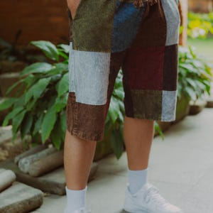 May include: Patchwork shorts in shades of brown, blue, green, and gray. The shorts are knee-length and have a drawstring waist. The person is wearing a white t-shirt, white socks, and white sneakers. The shorts are made of a patchwork design.