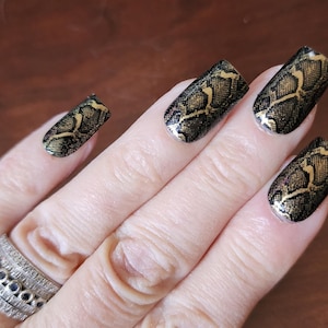 May include: Close-up of a hand with a silver ring and nails painted with a black and gold snakeskin pattern. The nails have a glossy finish, and the design is intricate and detailed.