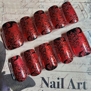 May include: Set of press-on nails with a red glitter base and black swirling design. The nails are rectangular shaped and displayed on a clear plastic holder. The words "Nail Art" are visible in the bottom right corner.