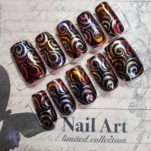 May include: A set of press-on nails with a black base and swirling patterns in red, orange, and silver. The nails are arranged on a clear display stand. The text "Nail Art limited collection" is visible.