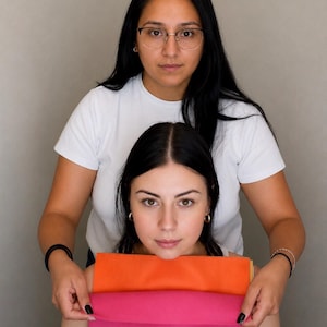 May include: A stack of rectangular fabric swatches in vibrant solid colors, including a bright orange top layer and a vivid hot pink bottom layer, held horizontally to display the texture and color saturation of the textile material.
