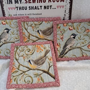 May include: Four square quilted fabric coasters with a bird and floral design. Each coaster features a light blue background with a bird perched on a branch with orange flowers. The edges are trimmed with a pink floral fabric. A sign in the background reads "IN MY SEWING ROOM THOU SHALT NOT..."