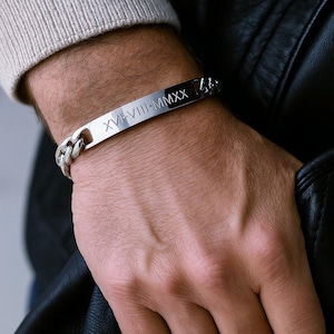 May include: A silver bracelet featuring a rectangular plate engraved with Roman numerals XVI-VIII-MMXX. The bracelet has a chunky chain link design. The image shows the bracelet on a person's wrist.
