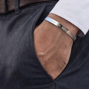 May include: A silver cuff bracelet with the engraved text "I LOVE YOU, WEIR". The bracelet is worn on a wrist, with the hand in a pocket. The cuff is a simple, elegant design.