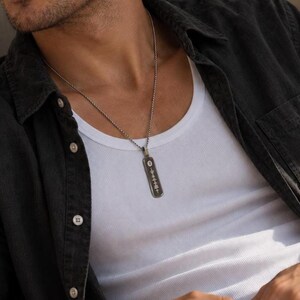 May include: A silver-toned metal pendant necklace featuring a rectangular tag with a vertical engraved sound wave pattern. The pendant hangs from a thin, textured silver-toned chain, resting against a white ribbed tank top and an unbuttoned dark shirt.