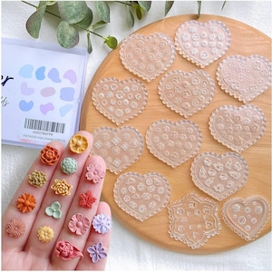 May include: A set of clear heart-shaped silicone molds with various floral and geometric textures, accompanied by a collection of small, colorful, molded clay flowers in shades of orange, yellow, pink, green, and purple, with a storage box.