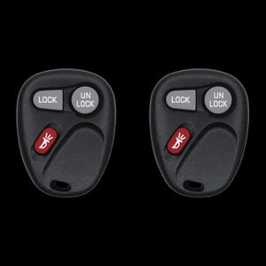 May include: Two black car key fobs with gray "LOCK" and "UN LOCK" buttons. Each fob has a red button with a horn symbol. The fobs are on a black background.
