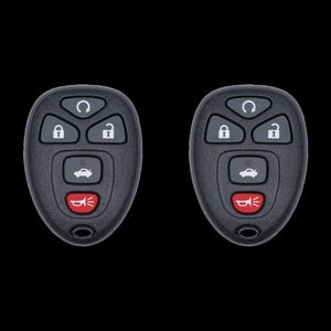 May include: Two black car key fobs with multiple buttons. The buttons include lock, unlock, panic, and a car icon. The panic button is red. The fobs are on a black background.