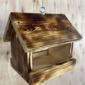 May include: A wooden bird feeder with a pitched roof and a rectangular feeding area. The wood has a natural grain pattern and a dark, burnt finish. A metal loop is attached for hanging.