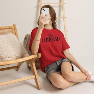 May include: A bright red short-sleeved t-shirt featuring the text "IN COMMAND" printed in bold black lettering across the chest. The shirt is paired with dark grey denim shorts, creating a casual look with a clear, prominent graphic design.