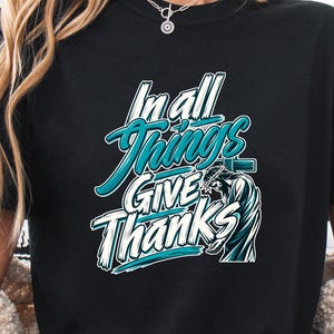 May include: A black short-sleeve t-shirt featuring the white and teal text "In all Things GIVE Thanks" with a graphic of a turkey. The shirt has a crew neckline and is paired with light blue denim shorts and a beige textured cardigan.