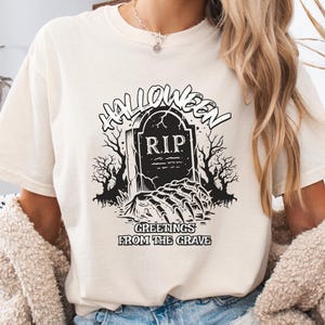 Może przedstawiać: Kremowy t-shirt z krótkim rękawem z czarnym nadrukiem graficznym przedstawiającym nagrobek z napisem R.I.P., otoczony nagimi drzewami i szkieletową dłonią wyłaniającą się z ziemi, z napisem HALLOWEEN GREETINGS FROM THE GRAVE.
