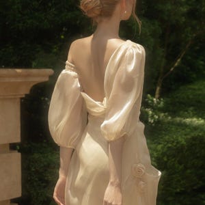May include: A cream-colored dress with a low back, featuring voluminous puff sleeves and a gathered bodice. The garment is crafted from a semi-sheer, shimmering fabric and includes two small, fabric-sculpted rose details on the side of the skirt.