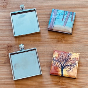May include: Four square pendant trays with silver-colored metal frames and bails. Two trays are empty, while the other two feature colorful, artistic designs. One has a winter forest scene, and the other has a tree design.