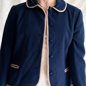 May include: A navy blue jacket with a rounded collar and light tan trim. The jacket has a button-up front and two small pockets with tan trim. The jacket is a classic style and appears to be made of a soft fabric.