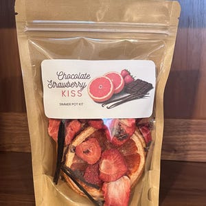 May include: A clear, resealable bag filled with a simmer pot kit. The kit includes dried strawberries, orange slices, and vanilla beans. A label reads "Chocolate Strawberry Kiss" with illustrations of fruit and chocolate.