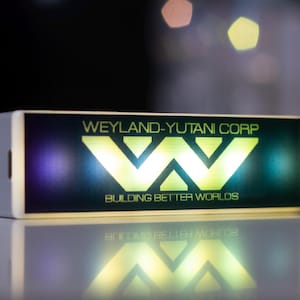 Op de afbeelding: Een rechthoekig, verlicht bord met de tekst "WEYLAND-YUTANI CORP" en het logo "W" in heldergroen. Daaronder staat "BUILDING BETTER WORLDS". Het bord staat tegen een donkere achtergrond.