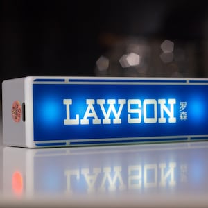 Op de afbeelding: Een rechthoekig, verlicht bord met het woord "LAWSON" in witte hoofdletters op een blauwe achtergrond. Het bord bevat ook Japanse karakters. De behuizing is wit met een blauwe rand en wordt weerspiegeld op een glanzend oppervlak.