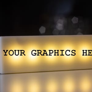 Puede incluir: Una caja de visualización rectangular, blanca e iluminada, con el texto negro "YOUR GRAPHICS HERE" en su cara frontal. El dispositivo emite un brillo cálido y suave desde el interior, resaltando las letras sobre la superficie opaca.