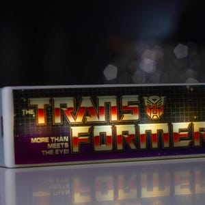 May include: A rectangular, white device featuring the Transformers logo and text. The logo is gold and red, with the words "THE TRANSFORMERS" and "MORE THAN MEETS THE EYE!" The device has a blue light on the side.