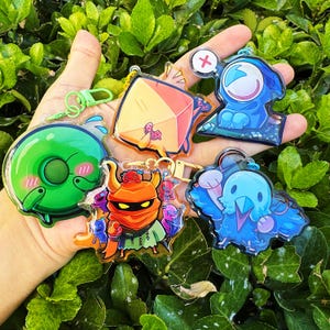 May include: A selection of vibrant acrylic keyrings showcasing cartoon characters. The keyrings include a green donut character, a blue bird, and a character with an orange mask. Each keyring is attached to a gold-coloured chain.