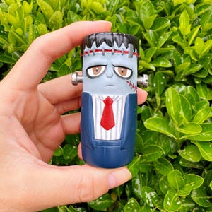 May include: A novelty lighter shaped like Frankenstein's monster. The lighter has a blue face, black hair, and a red tie. It is wearing a blue suit with silver bolts on the sides of its head. The background is a lush green bush.