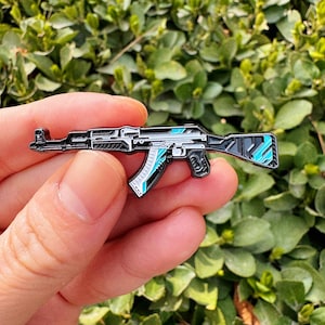 Puede incluir: Pin de esmalte detallado con forma de rifle de asalto AK-47. El pin presenta una combinación de colores negro, plateado y turquesa. El diseño incluye detalles intrincados de los componentes del arma.