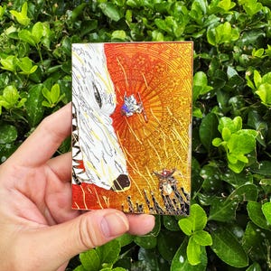 May include: A rectangular enamel pin featuring a stylized illustration. The design includes a white wolf's head, a small figure with a sword, and a detailed background in shades of orange and gold. The pin is held against a backdrop of green foliage.