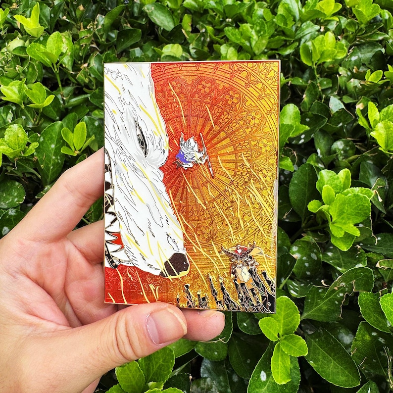 May include: A rectangular enamel pin featuring a stylized illustration. The design includes a white wolf's head, a small figure with a sword, and a detailed background in shades of orange and gold. The pin is held against a backdrop of green foliage.
