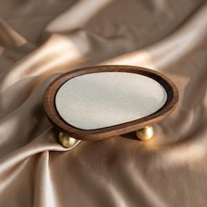 Könnte beinhalten: Ein Schmucktablett aus Holz mit einem hellfarbigen Stoffeinsatz. Das ovale Tablett wird von drei kleinen, goldfarbenen Kugelfüßen getragen. Das Holz hat eine satte, dunkelbraune Farbe, und der Hintergrund ist ein seidiger, goldener Stoff.