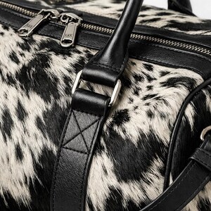 May include: A duffel bag featuring a black and white cowhide pattern with smooth black leather trim. The bag includes a top zip closure with silver-toned metal hardware, two sturdy leather handles, and a detachable shoulder strap with clips.