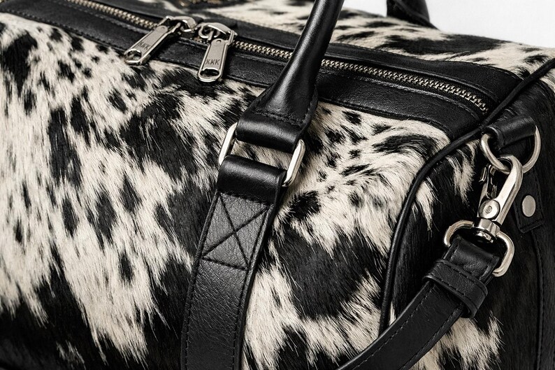 May include: A duffel bag featuring a black and white cowhide pattern with smooth black leather trim. The bag includes a top zip closure with silver-toned metal hardware, two sturdy leather handles, and a detachable shoulder strap with clips.