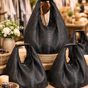 Peut inclure: Plusieurs sacs fourre-tout en cuir noir avec un motif peau de serpent sont présentés sur une table en bois. Les sacs, de tailles différentes, ont une seule poignée. L'arrière-plan comprend des vêtements et des objets décoratifs.