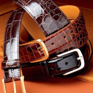 May include: Three leather belts in shades of brown and black are stacked on top of each other. The belts have a textured, crocodile-like pattern. The buckles are gold and silver. The belts are displayed on a brown surface.