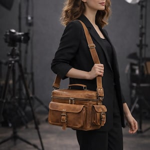 Puede incluir: Bolso mensajero de cuero marrón con múltiples bolsillos y una correa para el hombro. El bolso tiene forma rectangular con un asa superior y dos bolsillos delanteros con hebillas. El bolso lo lleva una persona con un traje negro.