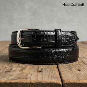 Puede incluir: Un cinturón negro con hebilla plateada, con textura de piel de cocodrilo. El cinturón se muestra sobre una superficie de madera. La marca "HosiCraftIntl" es visible en la esquina superior derecha.
