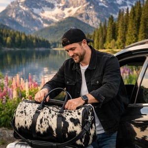 May include: A duffel bag featuring a black and white cowhide pattern with black leather handles, a detachable shoulder strap, and silver-toned metal hardware. The bag is positioned on top of a silver hard-shell suitcase with vertical grooves.