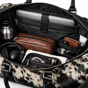 May include: A duffel bag with a black and white cow print pattern featuring black leather handles and trim. The interior is open, revealing a laptop compartment, a silver water bottle, a brown pouch, black headphones, and small organisers.