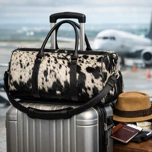 May include: A black and white cowhide duffel bag with black leather handles and a shoulder strap, resting on a silver metallic rolling suitcase. Beside the suitcase are a tan fedora hat, a brown passport holder, and black over-ear headphones.